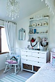 Romantic ornaments on chest of drawers and wall-mounted shelves and doll on rocking chair in white, child's bedroom
