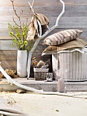 Various cushions and pouffe in shades of brown next to vase of flowers and branch against simple wooden wall