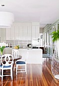 Wooden table and white-painted chairs in modern fitted kitchen with white-painted cupboards and ship deck flooring