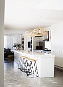 Barstools with metal frames at white, free-standing kitchen counter below wicker pendant lamps in designer apartment