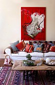 Bright red modern painting above country-house sofa with collection of cheerful cushions; coffee table upcycled from carved, repurposed wardrobe door