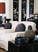 Bowl and antique vessel on black wooden trunk in front of armchairs with cushions; wall panels with Oriental-style floral patterns in background