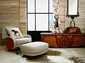 White armchair and matching footstool on animal-skin rug on floor next to rustic bench with leather seat cushion below window in wooden wall