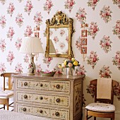 Table lamp with pleated fabric lampshade on painted chest of drawers below gilt-framed mirror on floral wallpaper