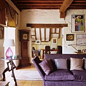 Purple sofa with scatter cushions next to side table in rustic interior with wood-beamed ceiling and view of dining area through wide open doorway