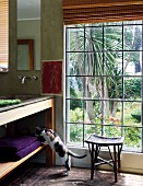 Rattan footstool in front of lattice window with view of garden and cat looking into base unit shelves of washstand