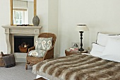 Felltagesdecke auf Doppelbett und Rattanlehnstuhl vor offenem klassischem Kamin im Schlafzimmer mit traditionellem Flair