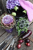 Globe thistles and artichoke flower arranged in ceramic pots next to aubergines, hydrangeas and pale violet cloth