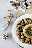 Festive table arrangement in natural shades with nuts on pale plates with silver rims, squares of chocolate on china board and white fir cones