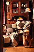 Candle lantern with knitted cover on tree trunk stool in front of comfortable armchair with knitted patchwork scatter cushions and antique dresser against wooden wall