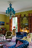 Traditional, English interior with strong colour contrasts between yellow patterned wallpaper, red chintz curtains, blue sofa and turquoise chandelier