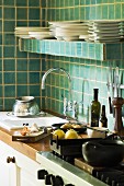 Corner of kitchen - kitchen counter with sink and cooker under shelf of crockery on green-tiled wall