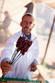 A shopkeeper demonstrating barbecued kebabs at a market (North Africa)