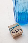 Small cardboard box decorated with blue rubber bands and lettering reading 'sorry' printed with hand-carved stamp