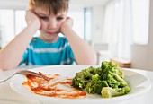 A boy sitting crossly in front of a plate of left-over vegetables