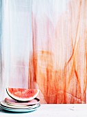 A slice of watermelon on a stack of plates in front of a transparent curtain