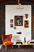 Orange-roter Sessel im Fifitesstil neben Couchtisch mit Tischleuchte vor weißem Paneel mit aufgehängten Bildern an Ziegelwand