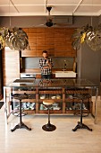Vintage barstools at counter with glass-fronted drawers in open-plan, fitted kitchen with pendant lamps with lampshades made from stylised leaves; man behind counter