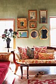 Scatter cushions with various patterns on antique bench in lounge-style room with collection of framed mirrors on green wall