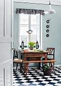 Antique, Biedermeier-style table and chairs below old hunting horn hanging in kitchen window; black and white gingham pelmet and chequered floor