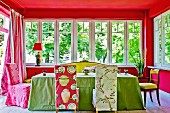 Colourful dining area in extension with bright, patterned loose chair covers and green tablecloth combined with red-painted walls