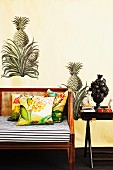 Partially visible bench with seat cushion and scatter cushions next to tray table against wall with painted pineapple motif
