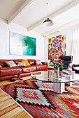 Round glass coffee table on rug with ethnic pattern in front of brown leather couch in living room with white, wood-beamed ceiling