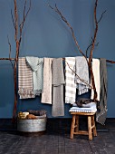 Scarves and blankets in pale, natural colours draped over frame made from long branches and stacked in zinc tub and on stool against blue wall