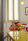 Arrangement of fruit in glass pedestal fruit bowl on yellow painted table with white base units and tall window in background