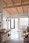 White, modern bathroom with wooden ceiling and free-standing bathtub below window next to rainfall shower and glass sliding door leading outdoors