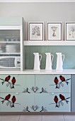 Silhouettes of birds on drawers of kitchen base unit; white china jugs on counter and framed drawings on shelf