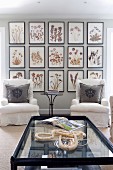 Coffee table with glass top and white armchairs in front of gallery of framed, floral pictures on wall