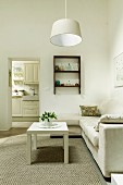 White coffee table on rug in front of pale sofa combination and simple pendant lamp in minimalist living room; doorway leading to kitchen in background