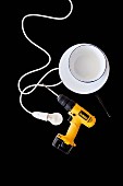 Yellow cordless drill, light bulb, electrical cable and white enamel bowls on black surface