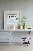 Moderne Kunst und Topfpflanzen in Körben auf schmalem Wandtisch mit Schubladenschrank vor grau marmorierter Wand; Hocker mit Schnurbespannung