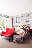 Roter Loungesessel und Sitzpolster vor Bett an Wand mit Ornamentmuster auf Tapete in modernem Schlafzimmer mit Retroflair