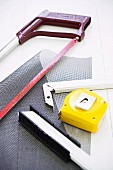 Red hacksaw, grey mesh, yellow tape measure and pieces of metal frame on white worksurface