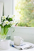 Breakfast for one - coffee cup on folded newspaper next to bouquet of white hyacinths in glass vase in front of vintage window