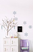 Festively decorated twigs in glass vase on white-painted cabinet against wall with bluish snowflake motifs and purple plexiglass chair with cushion