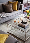 Small sculpture on stacked books and candles on glass and chrome coffee table; sofa, scatter cushions and rug in elegant shades of grey and brown