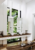 Floating washstand between two walls with twin countertop sinks and mirrors in front of window; rustic wooden bench in foreground