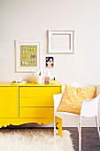 Gelbes Sideboard mit geschweiftem Sockel auf Flokatiteppich neben weißem Kunststoffstuhl, an der Wand weiße Bilderrahmen und ein Bild mit Frauenmotiv