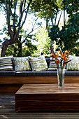 Vase mit Strelitzien auf Bodentisch aus tropischem Holz und Sitzbank mit Kissen auf Terrasse vor tropischem Garten