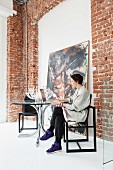 Woman sitting on designer chair at round glass table next to brick wall of loft interior with painting leaning on wall