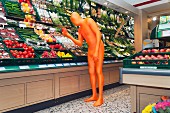A man in an orange costume looking at a pepper in the supermarket