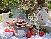 Gedeckter Teetisch mit Törtchen auf Platte, Äpfeln und Blumenstrauss im Freien