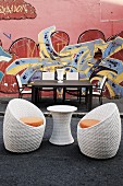 Gartentisch und Stühle sowie ein Tischchen aus Korbgeflecht mit zwei Schalenstühlen vor einer mit Graffiti bemalten Hauswand