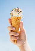 A hand holding an ice cream cone with a scoop of ice cream and colourful sugar sprinkles