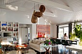 Open-plan interior with vintage ambiance and Long-Island style - lampshades made from natural materials hanging from white wooden ceiling and classic fifties chairs in dining area