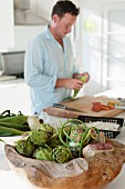 Artichokes and corn-on-the-cob in root wood bowl; man preparing meal on large chopping board in background